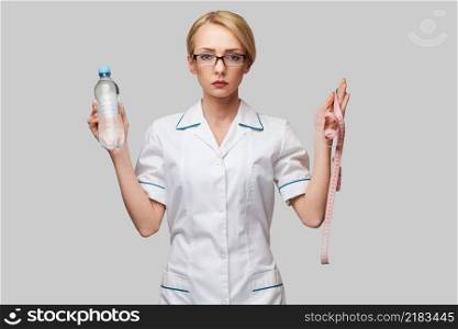 Portrait of young caucasian woman health care professional holding bottle of water and measuring tape standing over light grey background.. Portrait of young caucasian woman health care professional holding bottle of water and measuring tape standing over light grey background
