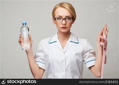 Portrait of young caucasian woman health care professional holding bottle of water and measuring tape standing over light grey background.. Portrait of young caucasian woman health care professional holding bottle of water and measuring tape standing over light grey background