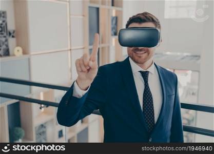 Portrait of young cheerful businessman in formal suit and VR headset goggles explores data visualized in virtual reality and touching it with index finger up in air while standing in company lobby. Young cheerful businessman in formal suit and VR headset goggles standing in company lobby