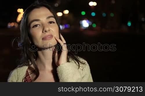 Portrait of young cheerful woman posing outdoors in the evening. Stunning long brown hair girl with enigmatic look and seductive smile flirting on city street at night against streetlights bokeh background. Slow motion. Steadicam stabilized shot.