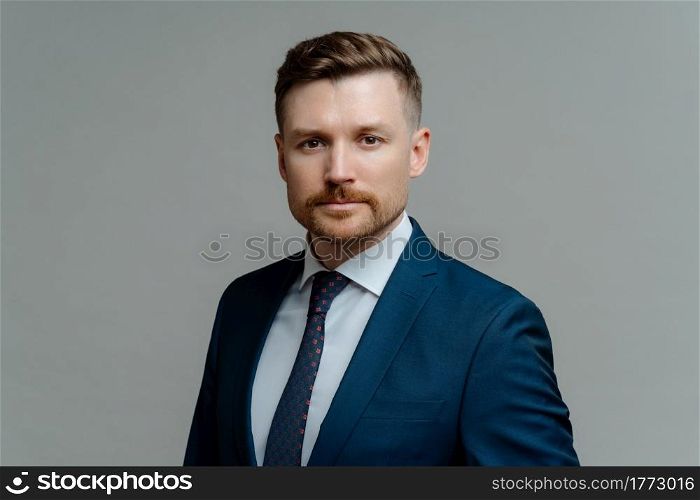 Portrait of young confident male entrepreneur or businessman wearing stylish suit looking at camera seriously while standing against grey background. Business people concept. Serious confident male boss or businessman in formal wear looking thoughtfully at camera