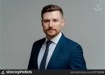 Portrait of young confident male entrepreneur or businessman wearing stylish suit looking at camera seriously while standing against grey background. Business people concept. Serious confident male boss or businessman in formal wear looking thoughtfully at camera
