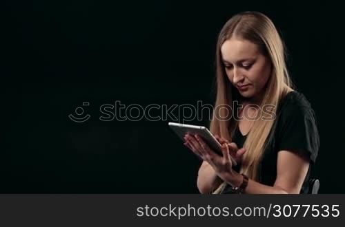Portrait of young confident woman surfing the net with touchpad isolated on black background. Beautiful female with long blonde hair working with digital tablet, scrolling webpage and searching online.