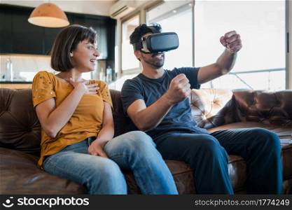 Portrait of young couple having fun together and playing video games with VR glasses while sitting on couch at home. New normal lifestyle concept.