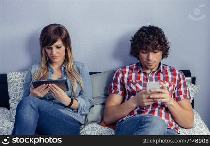 Portrait of young couple in love using electronic devices while resting over a bed. Leisure time at home concept.. Couple in love using electronic devices on bed