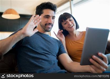Portrait of young couple on a video call with digital tablet while sitting on couch at home. Stay at home. New normal lifestyle concept.. Couple on a video call with digital tablet at home.