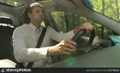 Portrait of young entrepreneur driving car on rural road during business trip in summer. Angle view. Confident business executive in necktie driving luxury car.