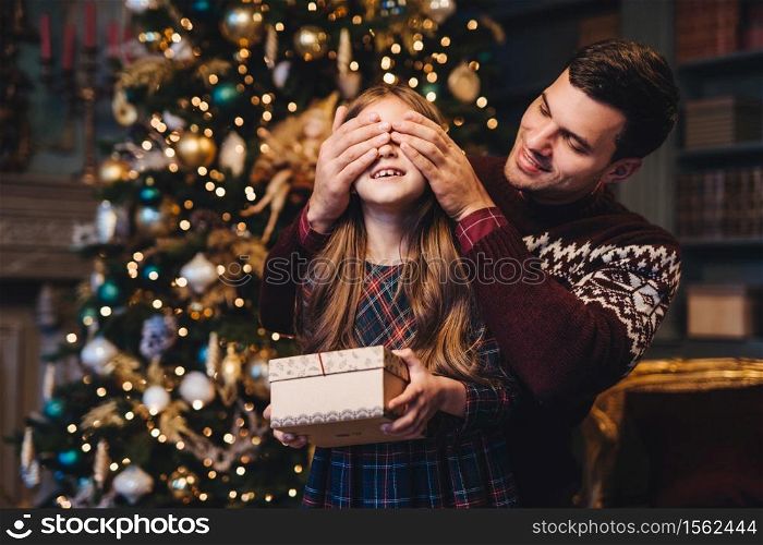 Portrait of young father covers his daughter`s eyes as going to make surprise for her, gives present, stand together near Christmas tree. Happy smiling girl recieves gift from dad. Surprise concept