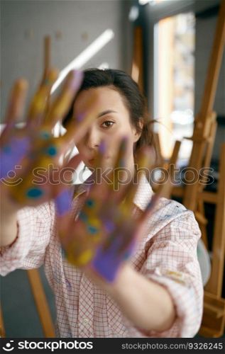 Portrait of young female creative artist peeping out of her painted hands looking at camera. University workshop and art studio class. Portrait of young female artist peeping out of her painted hands