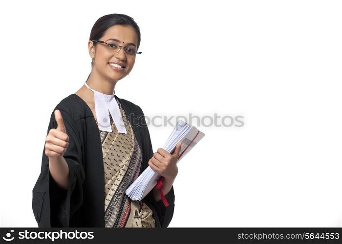 Portrait of young female lawyer showing thumbs up sign while holding documents against white background