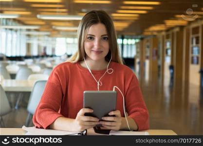 Portrait of young female student with earphones using digital tablet in the university library.