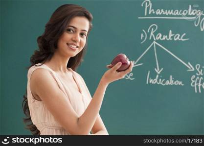 Portrait of young female teacher holding an apple against green board