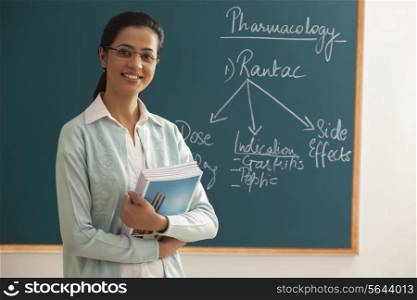 Portrait of young female teacher holding books against green board