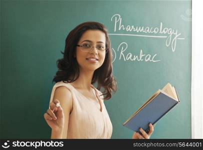 Portrait of young female teacher holding chalk and book against green board