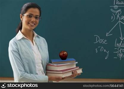 Portrait of young female teacher holding stack of books against chalkboard
