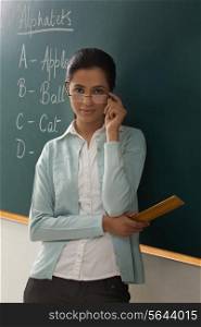 Portrait of young female teacher wearing glasses while leaning on chalkboard