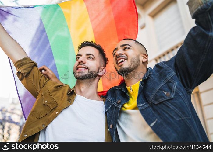 Portrait of young gay couple embracing and showing their love with rainbow flag at the street. LGBT and love concept.