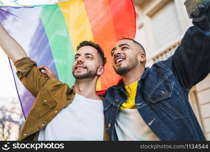 Portrait of young gay couple embracing and showing their love with rainbow flag at the street. LGBT and love concept.