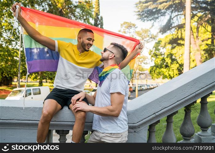 Portrait of young gay couple embracing and showing their love with rainbow flag in the stret. LGBT and love concept.