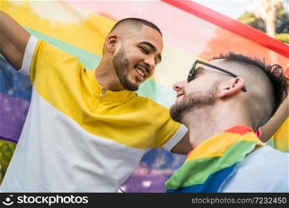 Portrait of young gay couple embracing and showing their love with rainbow flag in the stret. LGBT and love concept.