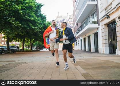 Portrait of young gay couple holding their hands and running together with rainbow flag in the street. LGBT and love concept.