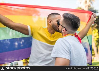 Portrait of young gay couple kissing and showing their love with rainbow flag in the stret. LGBT and love concept.