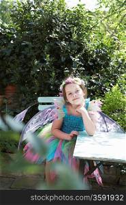 Portrait of young girl (5-6) in fairy costume sitting at garden table