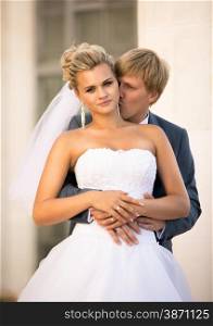 Portrait of young groom hugging blonde bride from back on street