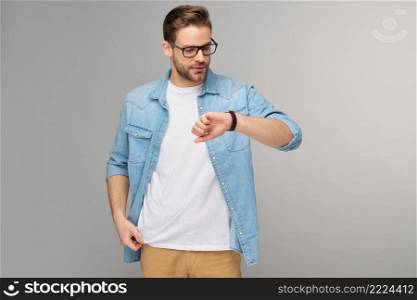 Portrait of young handsome caucasian man in jeans shirt looking at hand watch standing over light background.. Portrait of young handsome caucasian man in jeans shirt looking at hand watch standing over light background