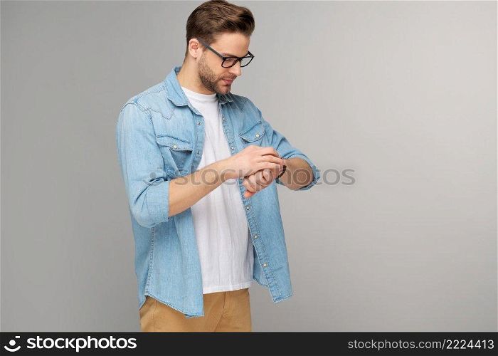 Portrait of young handsome caucasian man in jeans shirt looking at hand watch standing over light background.. Portrait of young handsome caucasian man in jeans shirt looking at hand watch standing over light background