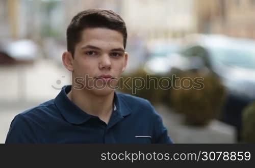 Portrait of young handsome man in casual polo-shirt smiling while sitting at street cafe against urbanscape background. Confident hipster teenager looking at camera with healthy toothy smile.