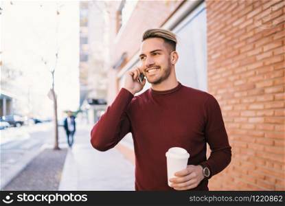 Portrait of young handsome man talking on the phone while holding a cup of coffee outdoors in the street. Communication concept.