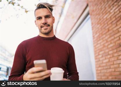 Portrait of young handsome man using his mobile phone while holding a cup of coffee outdoors in the street. Communication concept.