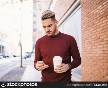 Portrait of young handsome man using his mobile phone while holding a cup of coffee outdoors in the street. Communication concept.