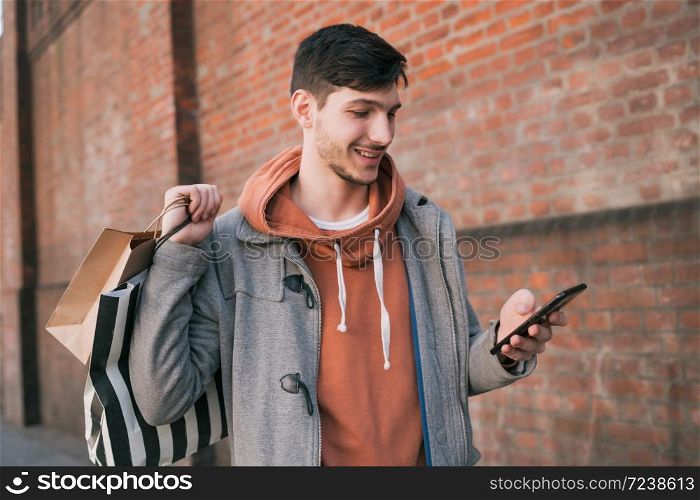 Portrait of young handsome man walking on the street, holding shopping bags and using his mobile phone. Urban, shop concept.