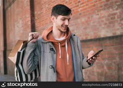Portrait of young handsome man walking on the street, holding shopping bags and using his mobile phone. Urban, shop concept.