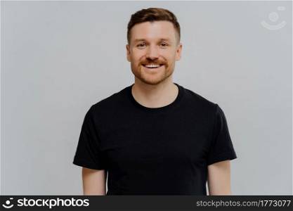 Portrait of young handsome smiling bearded man in black tshirt looking at camera with happy face expression, showing positivness while standing against grey background. Positive emotions concept. Young positive handsome man smiling at camera while posing against grey background