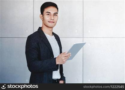 Portrait of Young Happy Businessman Using Digital Tablet. Standing by the Industrial Concrete Wall. Looking at Camera and Smiling