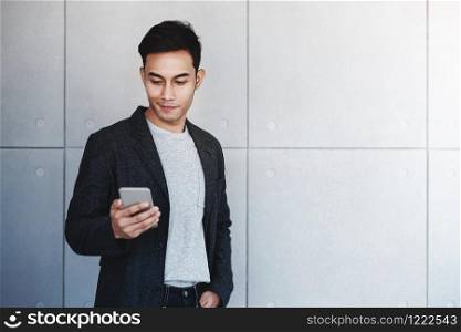 Portrait of Young Happy Businessman Using Smartphone. Standing by the Industrial Concrete Wall. Reading Message via Mobile Phone and Smiling