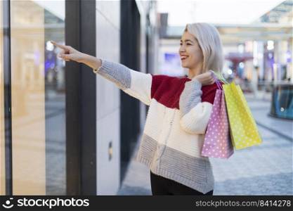 Portrait of young happy smiling woman with shopping bags pointing to a shopping mall in the street