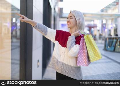 Portrait of young happy smiling woman with shopping bags pointing to a shopping mall in the street