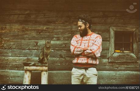 portrait of young hipster man with beard and cat in front of wooden house