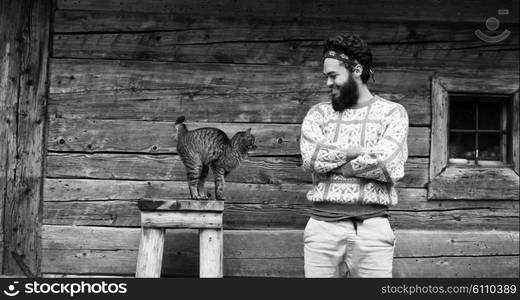 portrait of young hipster man with beard and cat in front of wooden house