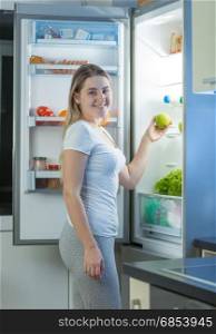 Portrait of young hungry woman taking apple from refrigerator at night