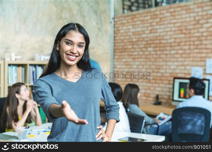 Portrait of young indian trangender extending hand and standing in creative workplace office with team work colleagues working and meeting in backgound. Welcome to my team scene and concept.