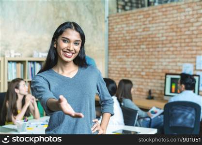 Portrait of young indian trangender extending hand and standing in creative workplace office with team work colleagues working and meeting in backgound. Welcome to my team scene and concept.