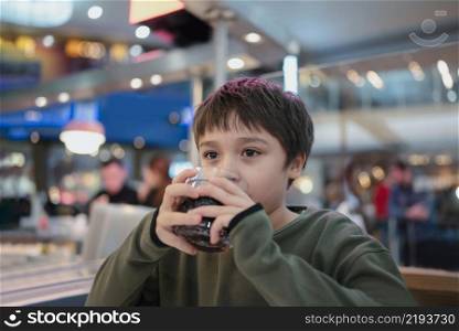Portrait of Young kid sitting on table drinking cold drink in restaurant, A boy drinking soda or soft drink from glass, Child boy waiting for food in cafe