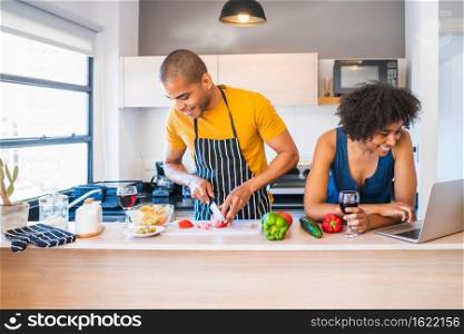 Portrait of young latin couple using a laptop while cooking in kitchen at home. Relationship, cook and lifestyle concept.
