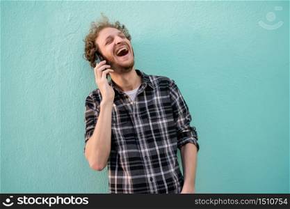 Portrait of young latin man laughing and talking on the phone against light blue background. Communication concept.