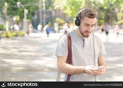 Portrait of young latin man typing on his phone with black headphones. Outdoors.